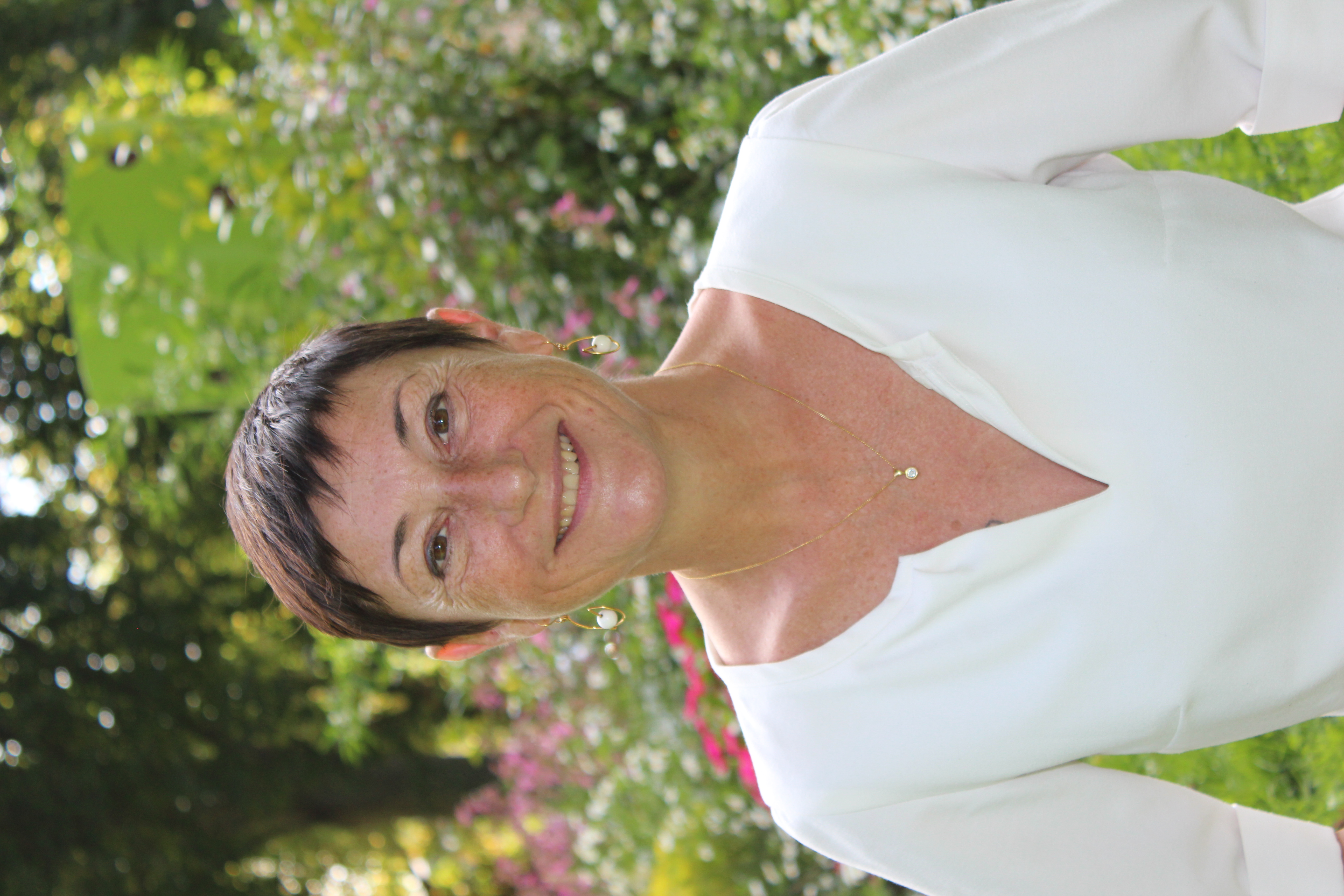 photo de la thérapeuthe. une femme, cheuveux courts et bruns, yeux bruns, qui sourie, vêtue d'un T-shirt blanc. 
              En arrière plan il y a des fleures.